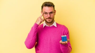 Young caucasian man holding Rubik’s cube isolated on yellow background pointing to temple with finger, thinking.