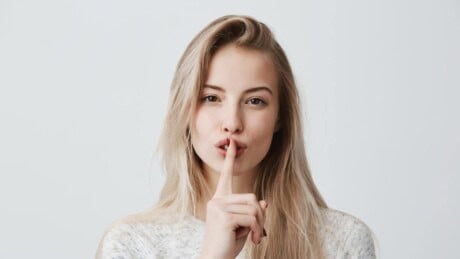 Shot of beautiful female with long blonde hair showing hush sign with a serious expression.