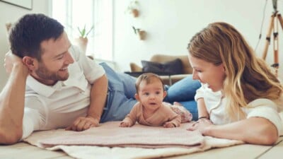 A young mom and dad laying on a blanket on the floor with their baby smiling at eachother.