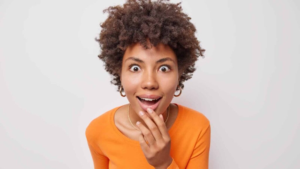 African American woman looking intrigued keeps mouth widely opened dressed in casual orange shirt isolated over gray background