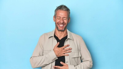 Middle-aged caucasian man on blue backdrop laughs happily and has fun keeping hands on stomach.