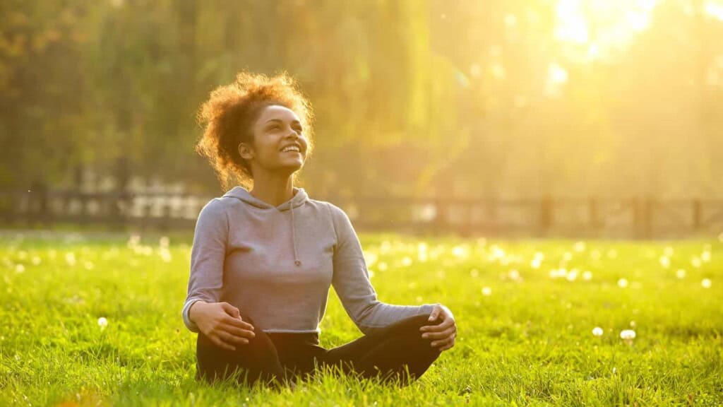 Woman sitting indian style in the grass, smiling while looking up, with the sunlight at her back