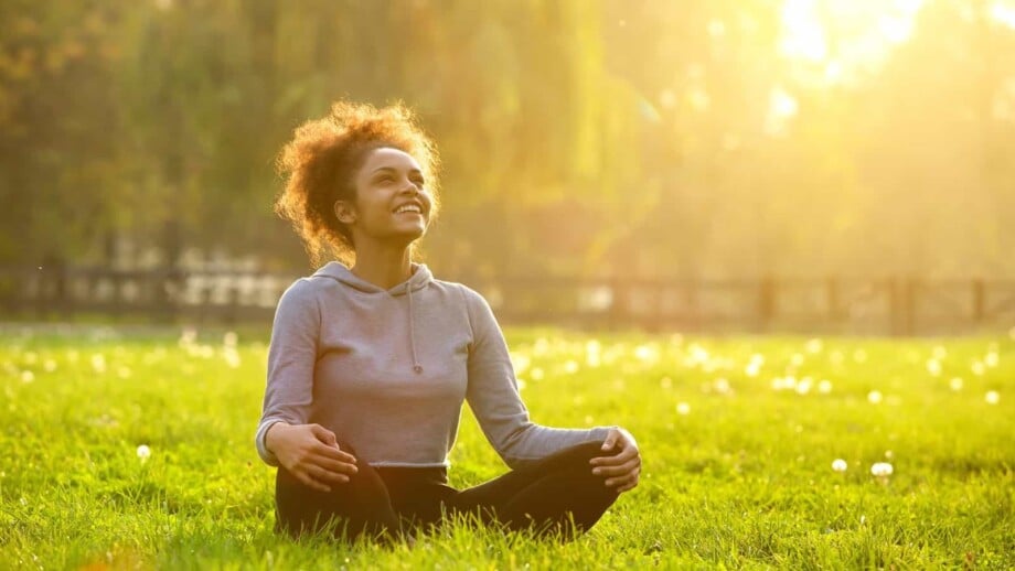 Woman sitting indian style in the grass, smiling while looking up, with the sunlight at her back